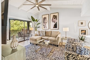 A living room with a sofa, coffee table, and patterned rug. There are two framed pictures on the wall, a ceiling fan, and a plant. - The Ricardo on Ramon (Palm Springs)