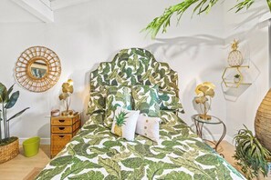 Ein Bett mit einem Kopfende mit Blattmuster, ein runder Spiegel und ein kleiner Nachttisch mit einer Ananas-Dekoration.
