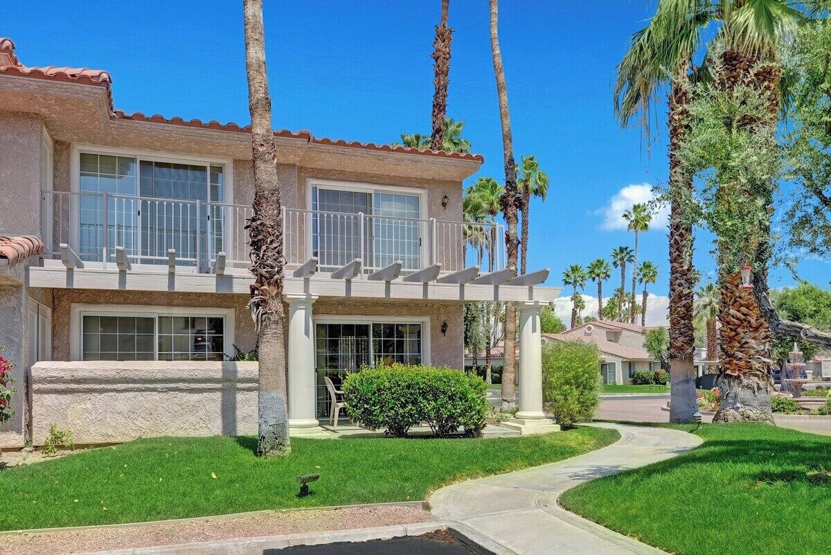 A two-story house with a balcony, surrounded by palm trees and a well-maintained lawn.