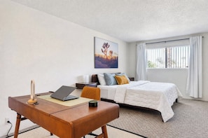 A hotel room with a bed, a desk with a laptop, a chair, a painting on the wall, and a window with curtains. - Casa Palm Canyon - South PS (Palm Springs)