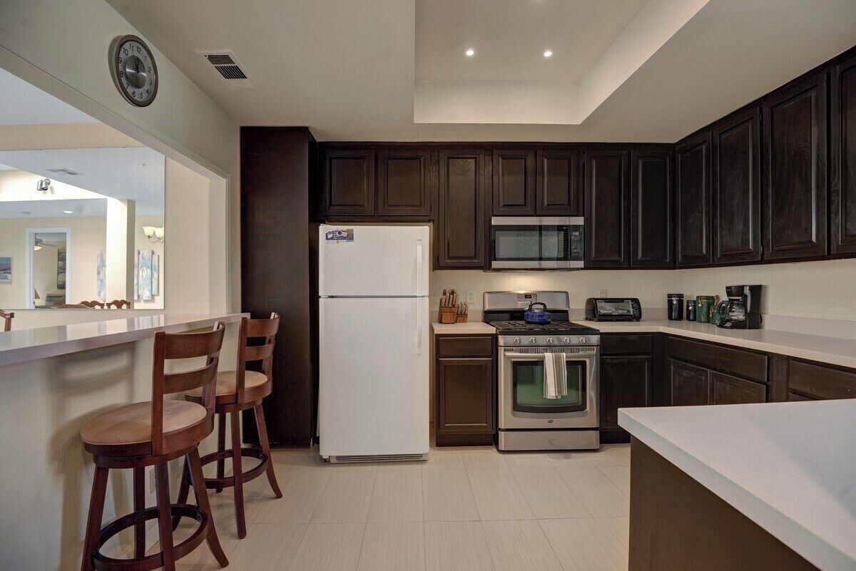 A kitchen with dark wooden cabinets, a white refrigerator, a microwave, an oven, and a bar area with two wooden stools.