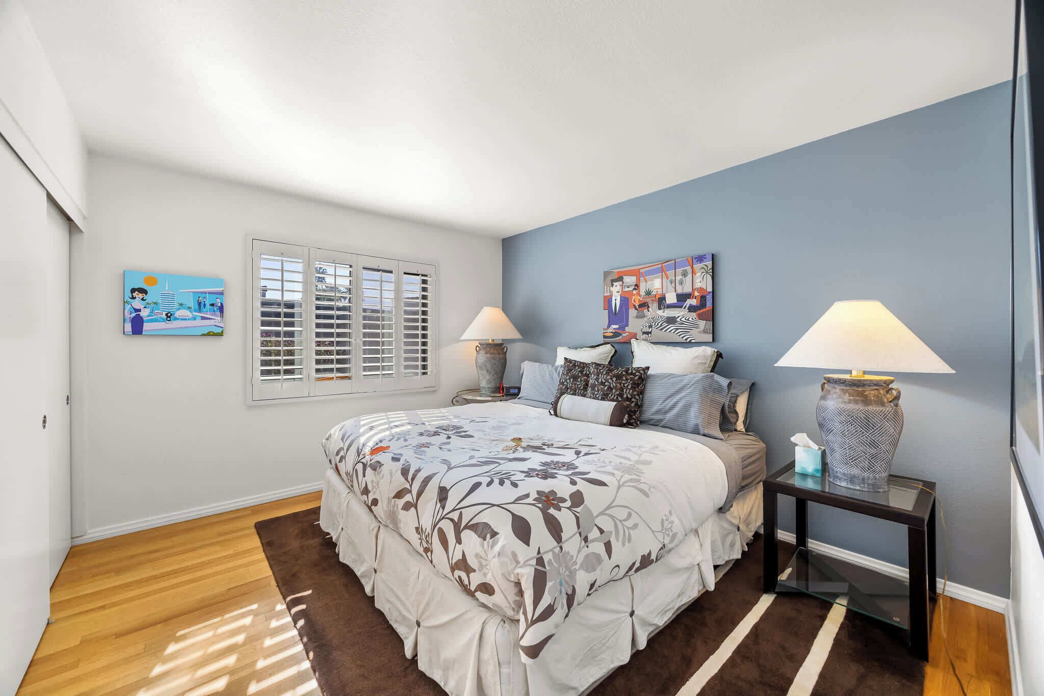 A bedroom with a bed, two bedside tables, a window with blinds, and artwork on the walls.