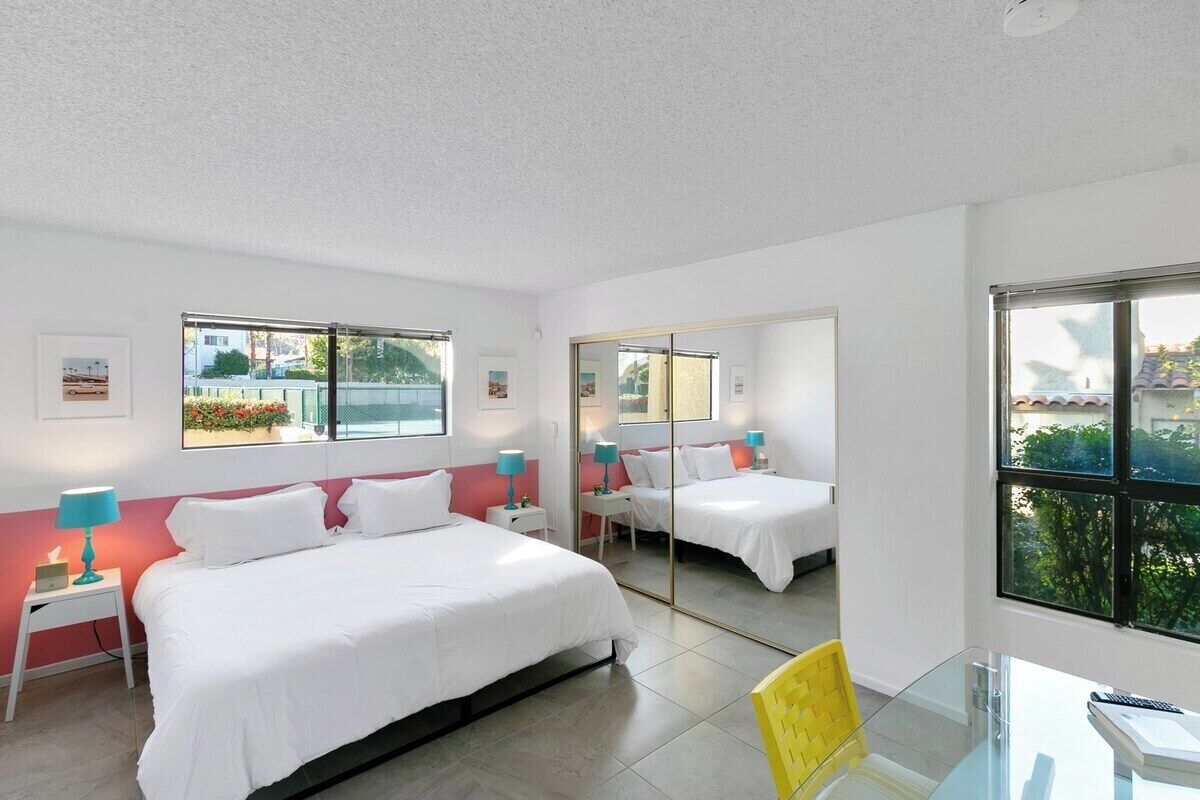 A hotel room with two beds, a desk, and a chair. There are windows with views of greenery and buildings.