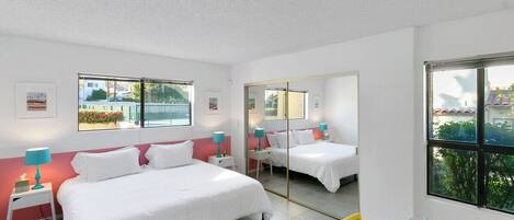 A hotel room with two beds, a desk, and a chair. There are windows with views of greenery and buildings.