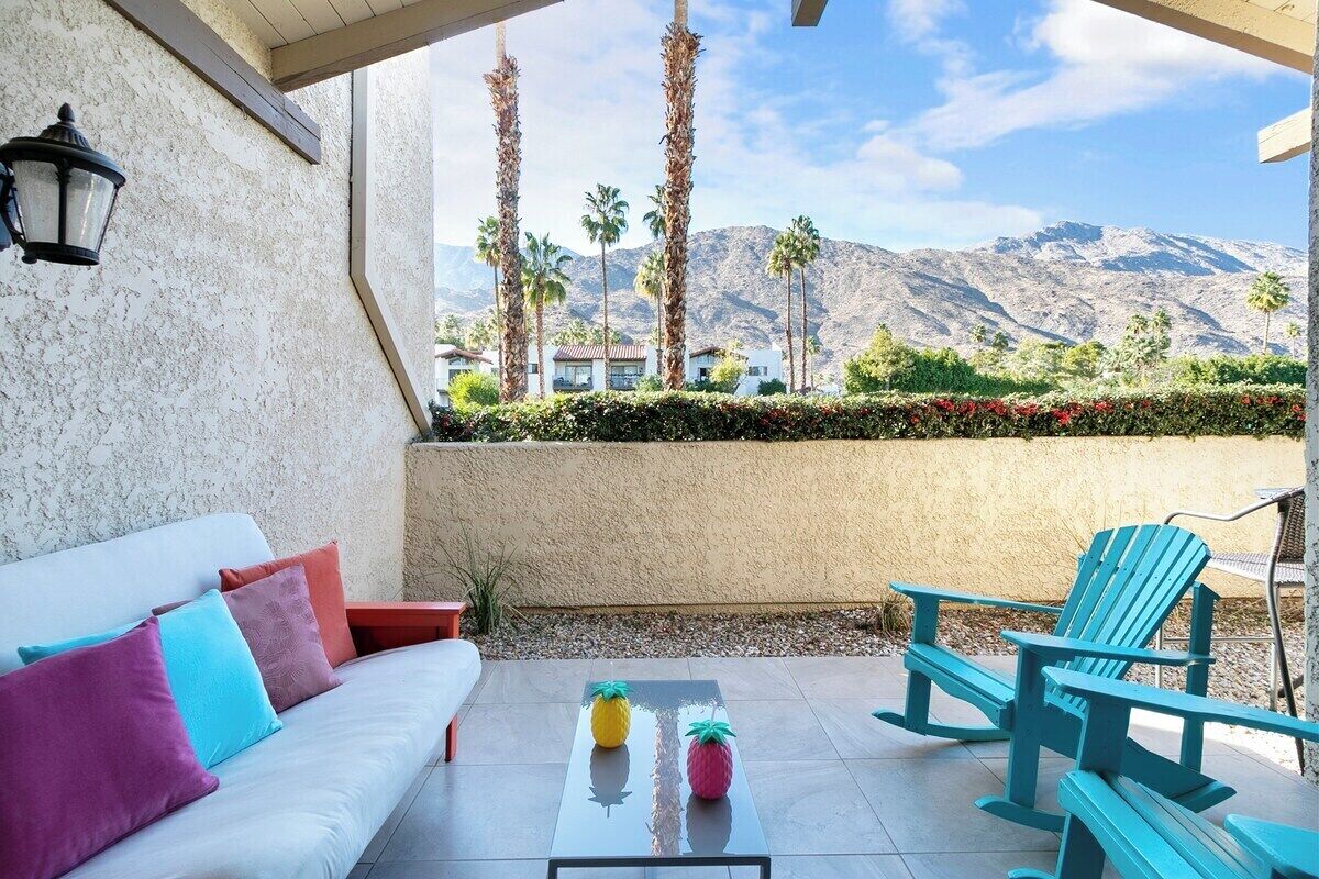 A patio with a white sofa, colorful cushions, a glass table with two pineapple-shaped objects, and teal Adirondack chairs.
