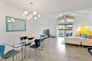 A modern living room with a glass dining table, black chairs, a sofa with colorful cushions, a television, and a large window. - Canyon Country Club Chic (Palm Springs)
