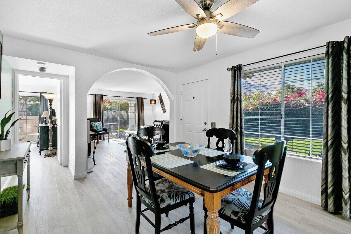 Comedor con mesa para cuatro, ventilador de techo y vista a través de una ventana con cortinas.