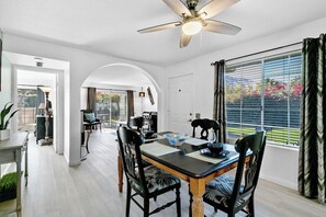 A dining area with a table set for four, a ceiling fan, and a view through a window with curtains. - Mesquite Delight - 2BR Condo with Mountain Views (Palm Springs)