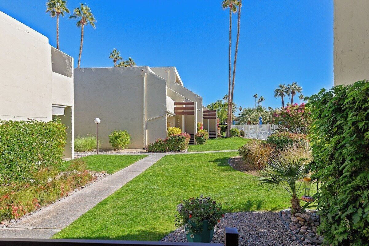 A residential area with a pathway leading to a building, surrounded by greenery and palm trees.