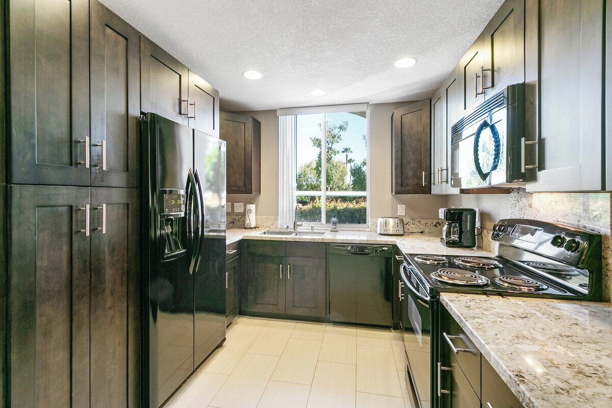 A modern kitchen with dark cabinetry, a stainless steel refrigerator, a built-in oven, and a large window providing natural light.