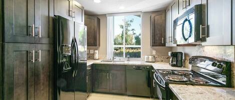 A modern kitchen with dark cabinetry, a stainless steel refrigerator, a built-in oven, and a large window providing natural light.