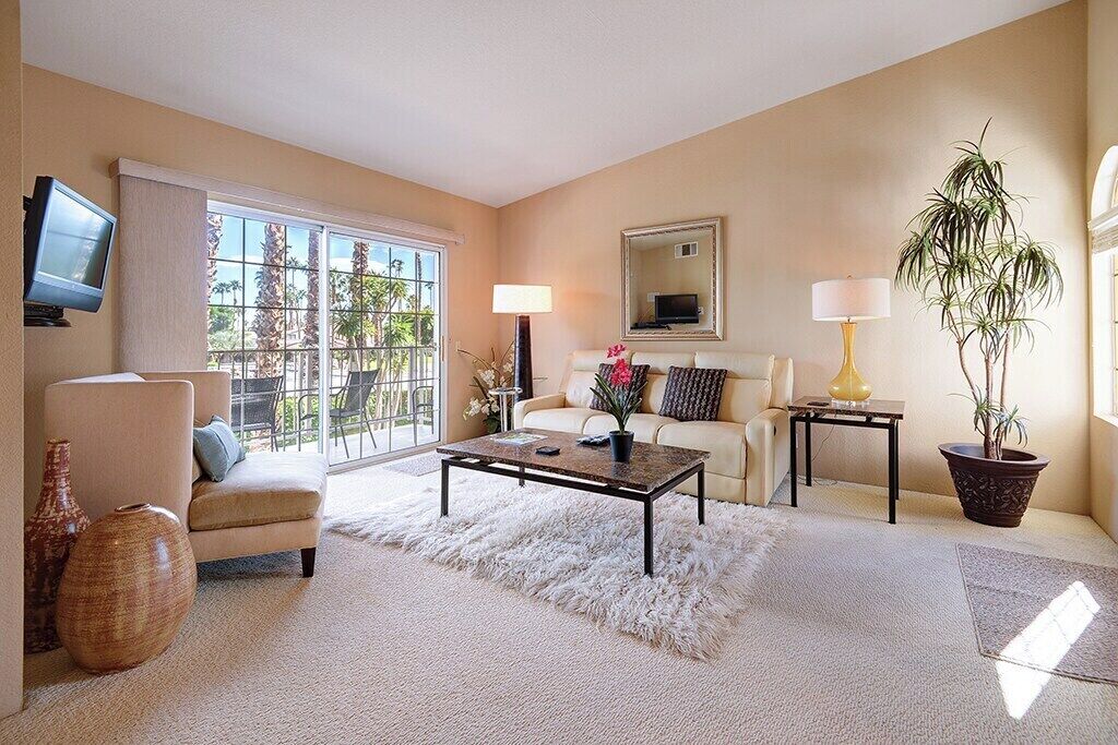 Ein Wohnzimmer mit Sofa, Sessel, Couchtisch, Fernseher und einem großen Fenster mit Blick auf Palmen.
