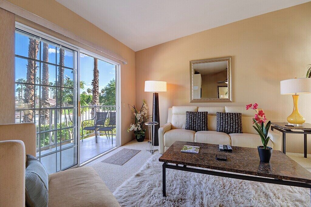 A living room with a sofa, coffee table, and a large window with a view of palm trees.