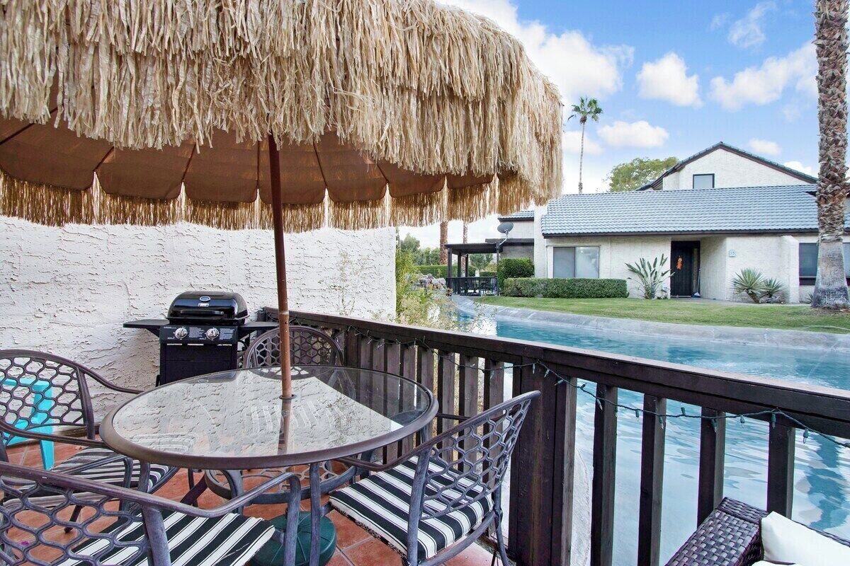 Um pátio com mesa e cadeiras redondas, churrasqueira e piscina ao fundo.