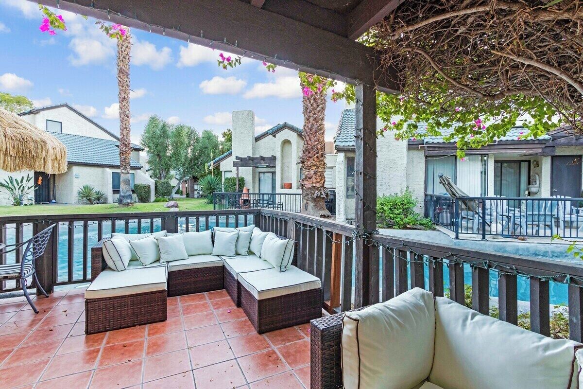 A covered outdoor seating area with a pool view, a thatched-roof building, and a modern house in the background.