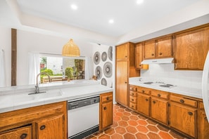 A kitchen with wooden cabinets, a white countertop, a built-in dishwasher, and a large window with a view of the outdoors. - Araby Luxe by Design (Palm Springs)