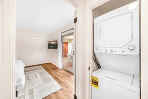 A laundry room with a white washing machine, a white door leading to a balcony, and a bed with a white cover in the adjacent room. - Araby Luxe by Design (Palm Springs)