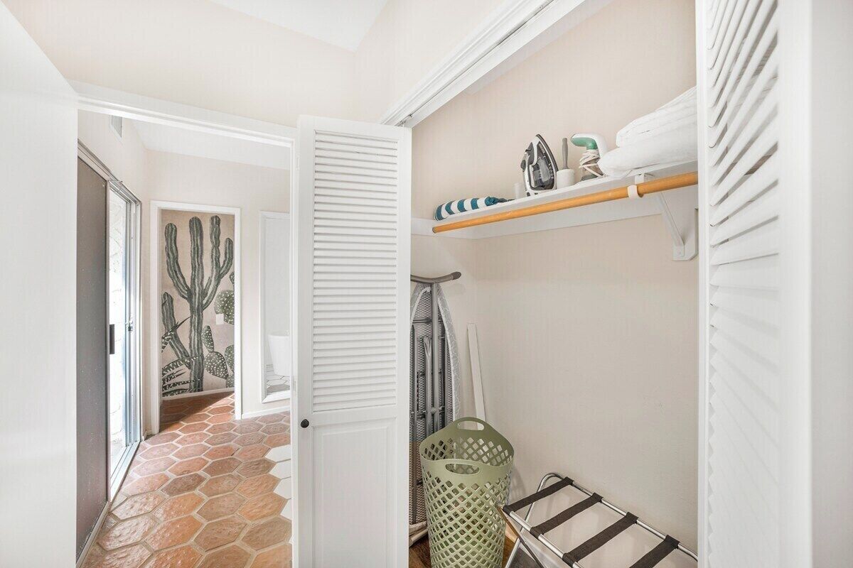 Un pasillo con piso de baldosas, una puerta, un cesto de ropa sucia y un estante con productos de limpieza.