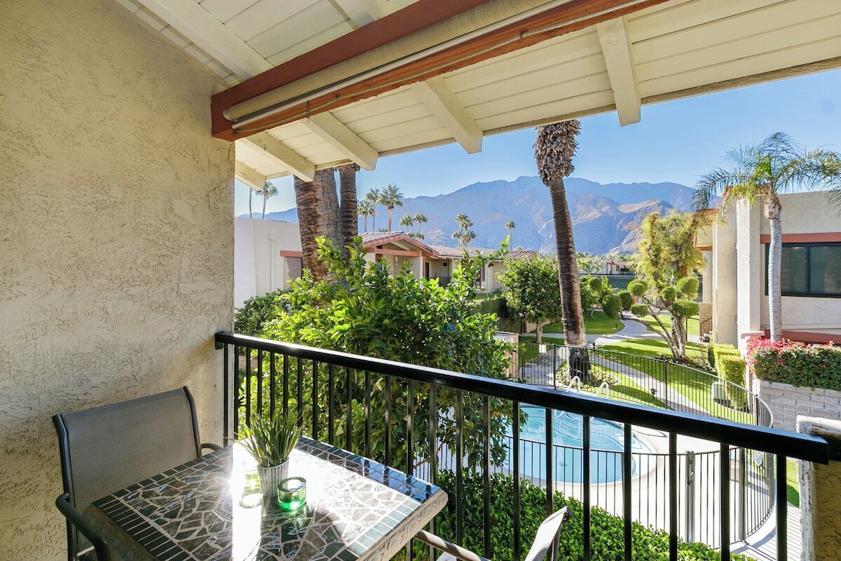 Un balcon avec une table et des chaises, donnant sur une piscine et un espace paysager agrémenté de palmiers, avec une chaîne de montagnes en arrière-plan.