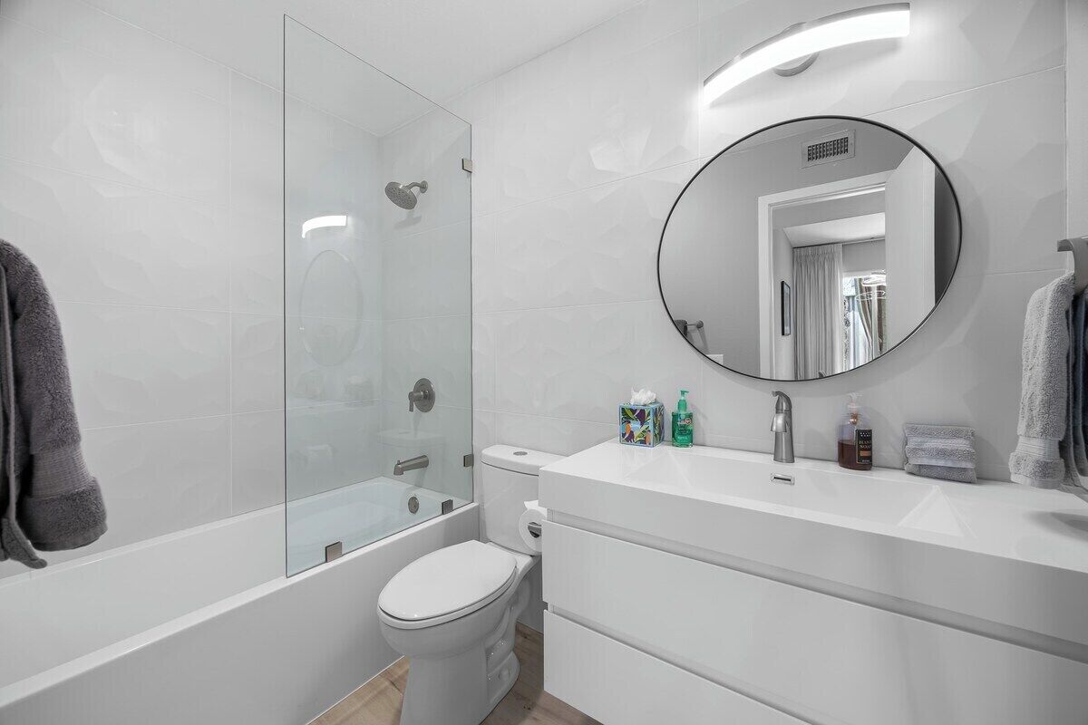 Une salle de bain moderne avec une douche à parois de verre, des toilettes blanches et un meuble-lavabo agrémenté d’un miroir rond.