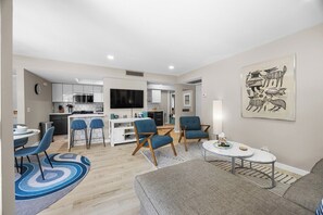 Un salon moderne avec un canapé gris, une table basse et des chaises bleues. On aperçoit une cuisine à l’arrière-plan, avec des meubles blancs et un téléviseur à écran plat.