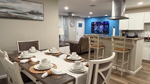 A dining area with a table set for four, a kitchen in the background, and a living room with a TV. - 4 Bedroom Coastal Farmhouse In Cocoa Florida (Cocoa)