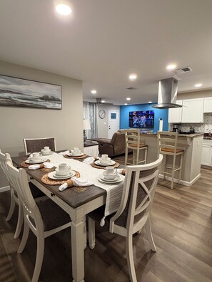 A dining area with a table set for four, a kitchen in the background, and a living room with a TV. - 4 Bedroom Coastal Farmhouse In Cocoa Florida (Cocoa)