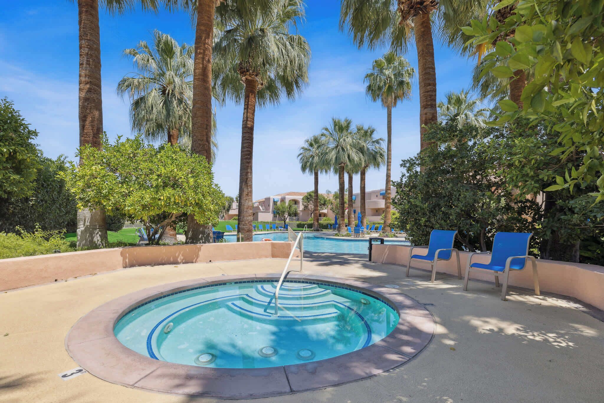 A swimming pool with a hot tub, surrounded by palm trees and blue lounge chairs.