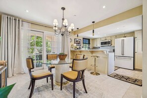 A modern kitchen with a central island, a dining area with a glass table and chairs, and a view of greenery outside. - Deauville Amora - Downtown PS (Palm Springs)