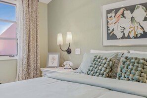 A hotel room with a bed, a nightstand, a lamp, a framed picture, and a window with curtains. - Deauville Amora - Downtown PS (Palm Springs)