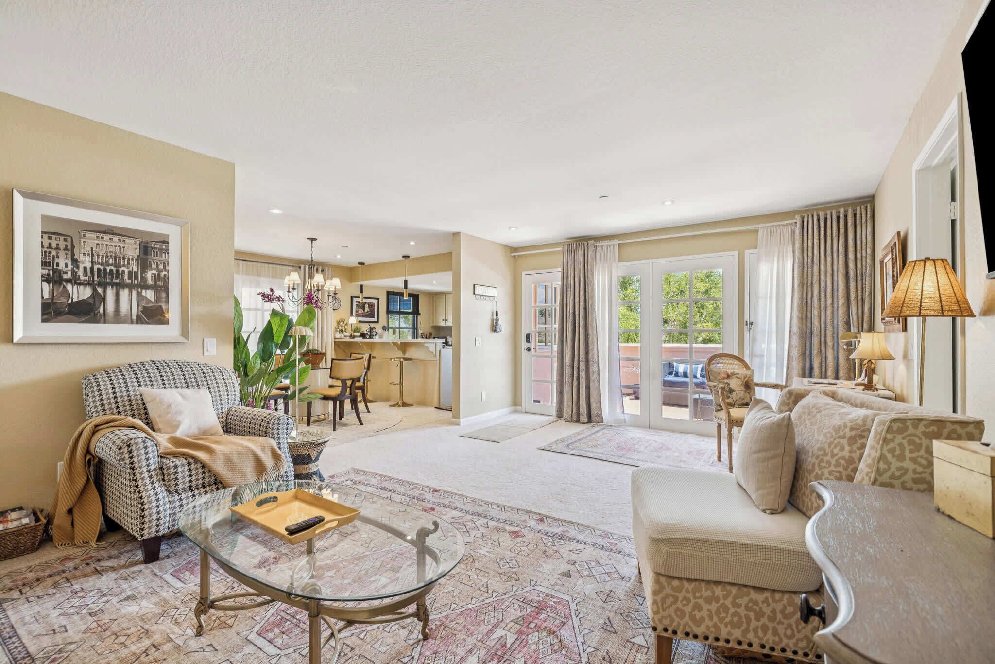 A spacious living room with a sofa, armchair, and a glass coffee table. There is a dining area with a table and chairs, and a kitchen in the background.