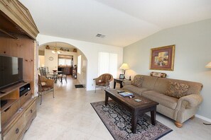 A living room with a sofa, coffee table, and television. There is a dining area with chairs and a table in the background. - Mesquite Tranquility Phase 3 (Palm Springs)