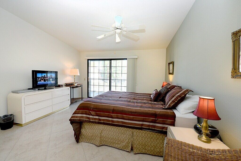 A bedroom with a bed, a television, a ceiling fan, a window, and a bedside table with a lamp.