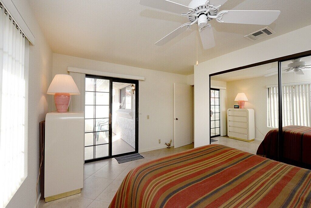 Habitación de hotel con cama, ventilador de techo, ventana con persianas, puerta que da a un balcón y cómoda con lámpara.