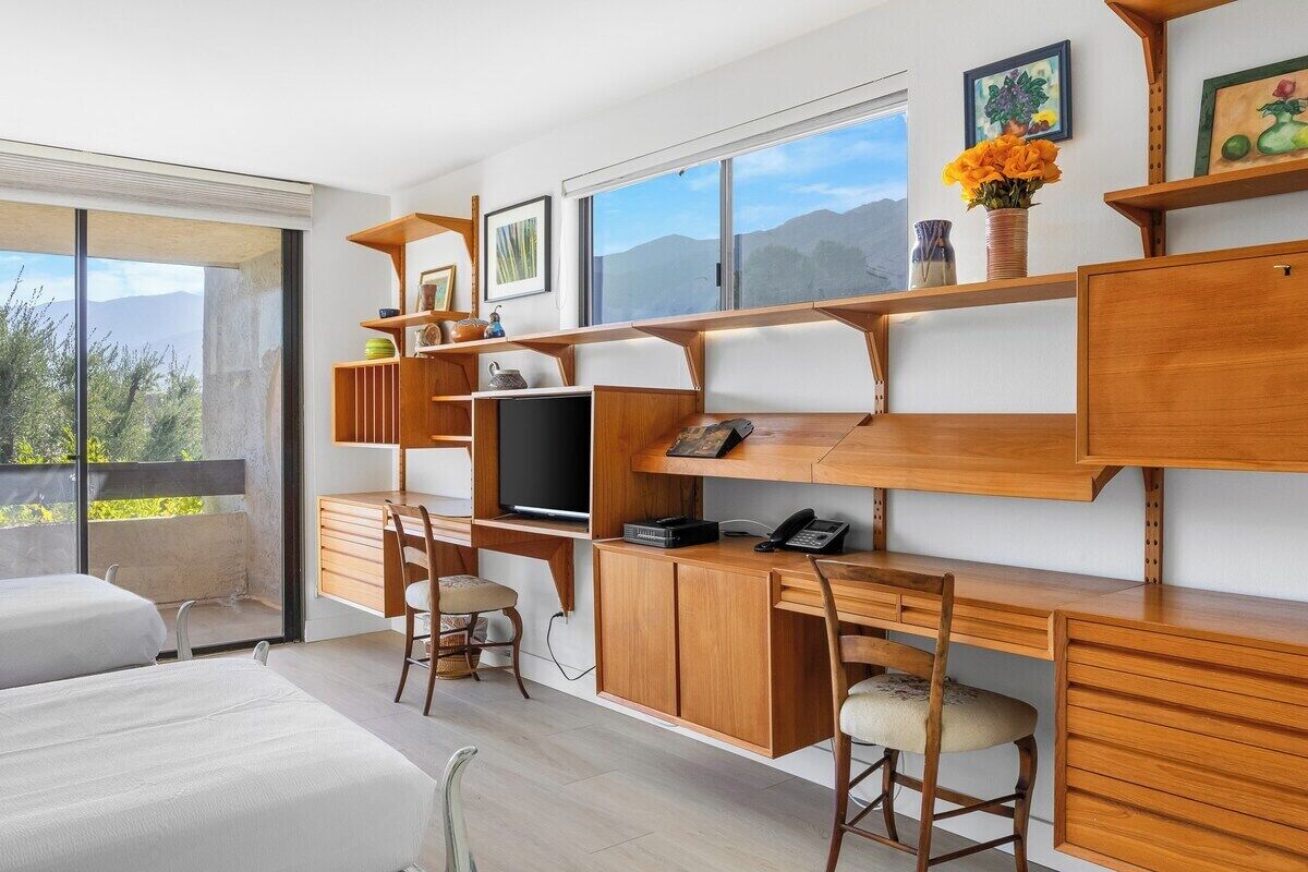 Ein modernes Schlafzimmer mit einem großen Bett, einem Schreibtisch und einem Sessel. Ein Fernseher und ein Regal mit Dekorationselementen sind ebenfalls vorhanden.