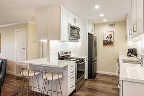 A modern kitchen with white cabinetry, stainless steel appliances, and a bar area with stools. - Ramon Estados Retreat (Palm Springs)