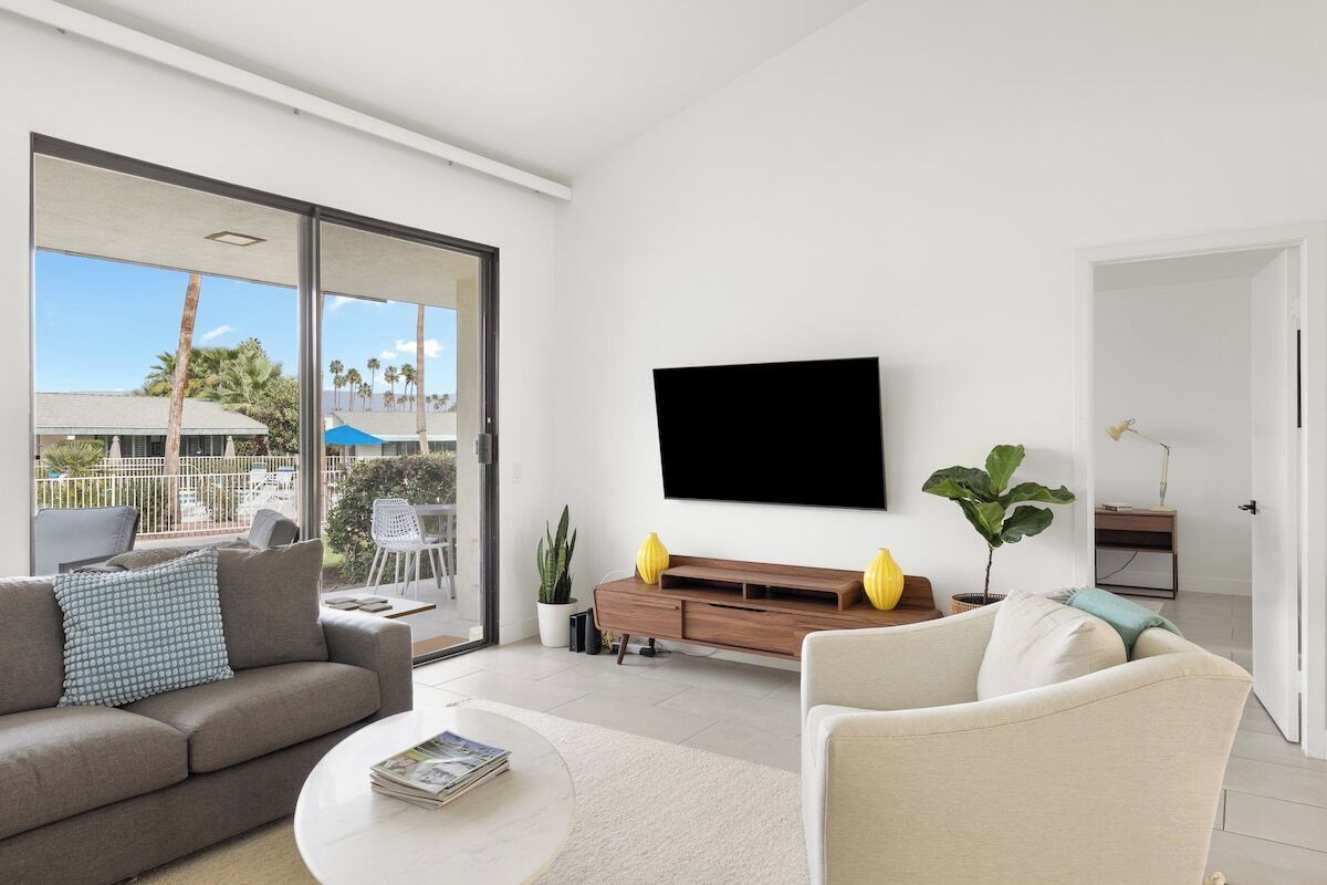 A modern living room with a flat-screen TV mounted on the wall, a sofa, a coffee table, and a sliding glass door leading to an outdoor area.