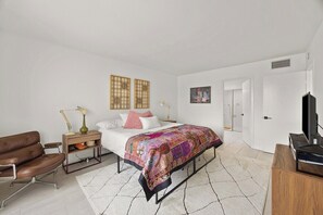 Quarto moderno com cama, mesas de cabeceira, escrivaninha, cadeira, televisão e porta.