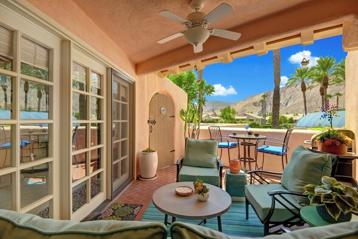 A sunlit patio with a ceiling fan, outdoor seating, and a view of palm trees and mountains.