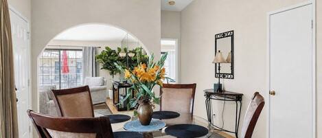 Een eethoek met een glazen tafel, stoelen, een vaas met bloemen en een spiegel aan de muur.