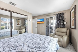 Ein Schlafzimmer mit einem Bett, zwei Stühlen, einem Fernseher und einer Schiebetür, die auf einen Balkon führt.