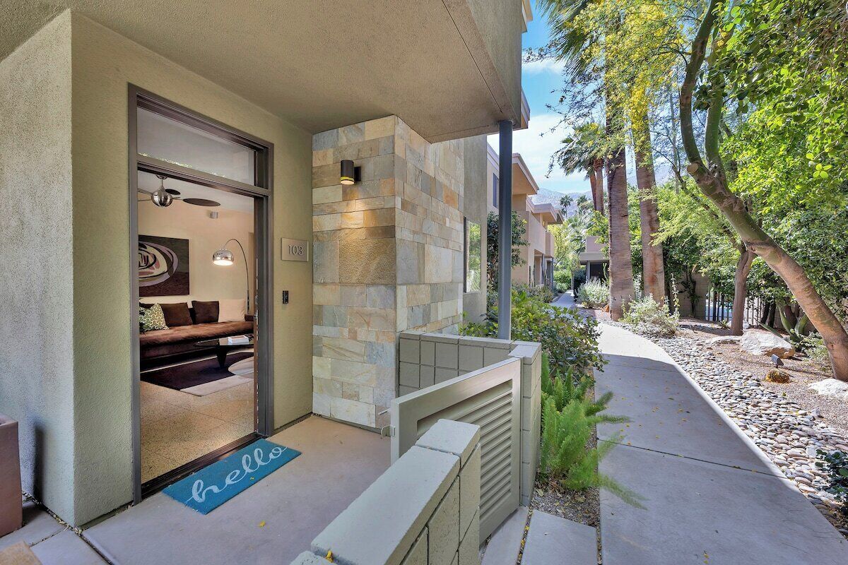 Ingresso moderno di un edificio con facciata in pietra, un portico coperto e un vialetto che conduce a una zona relax con divano e tavolino.