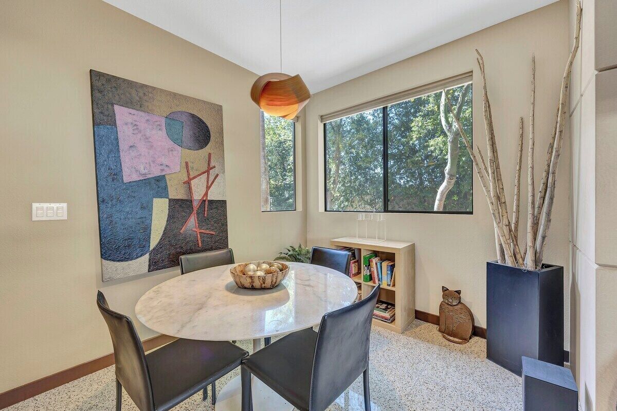 A dining area with a round table, chairs, a bowl of fruit, a bookshelf, and a window with a view of trees.