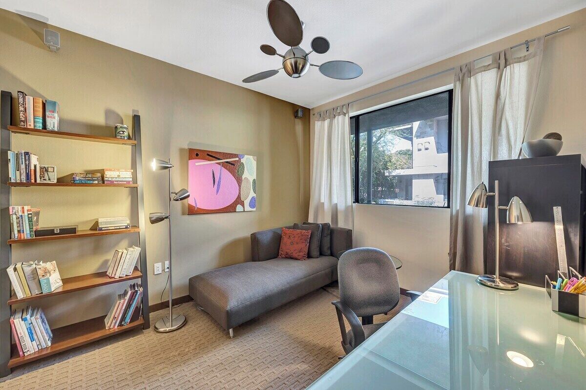 Une pièce comprenant un canapé, un bureau, une bibliothèque, un ventilateur de plafond et une fenêtre avec des rideaux.
