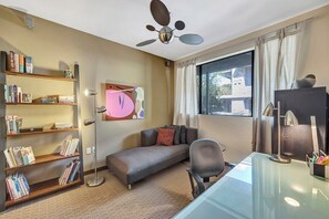 Une pièce comprenant un canapé, un bureau, une bibliothèque, un ventilateur de plafond et une fenêtre avec des rideaux.
