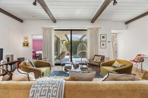 A living room with a sofa, chairs, a coffee table, and a bookshelf. There is a sliding glass door leading to an outdoor area with a pool and greenery. - Charming single-story condo boasts three bedrooms (Palm Springs)
