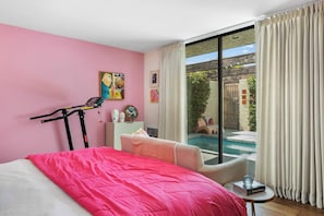A bedroom with a pink wall, a bed with a pink blanket, a treadmill, and a view of a pool and outdoor area. - Charming single-story condo boasts three bedrooms (Palm Springs)
