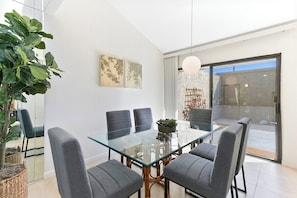 A modern dining area with a glass table, grey upholstered chairs, and a large potted plant. - Saddlerock Garden Haven (Palm Springs)