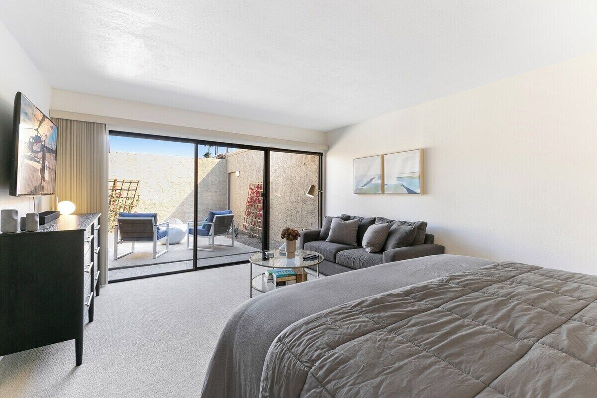 A modern hotel room with a bed, a sofa, a desk, and a balcony with outdoor seating.