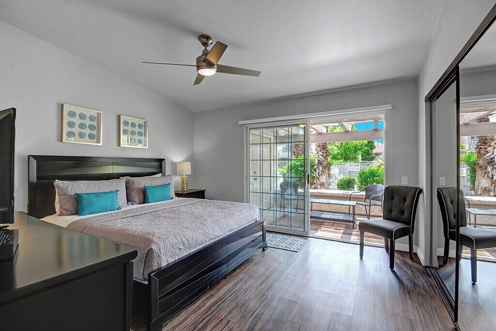 A bedroom with a bed, a desk, a chair, a ceiling fan, and a sliding glass door leading to an outdoor area.